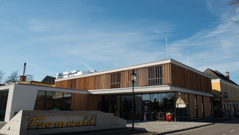 Der Hubertushof in Bad Fischau-Brunn, © Fotografin: Eva Heindl Modernes Gebäude mit Holzfassade und Glasfront, davor ein Schild mit der Aufschrift 'Fromwald'.