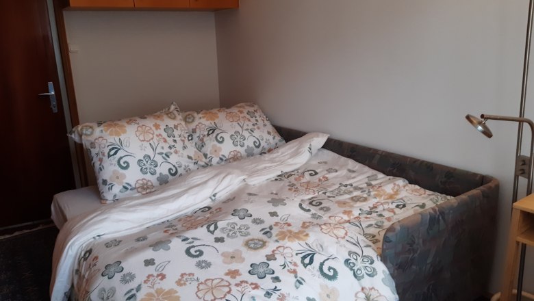 Ein kleines Zimmer mit einem ausziehbaren Sofa, das als Bett f&uuml;r zwei Personen dient, mit floraler Bettw&auml;sche und einem Leselicht daneben.