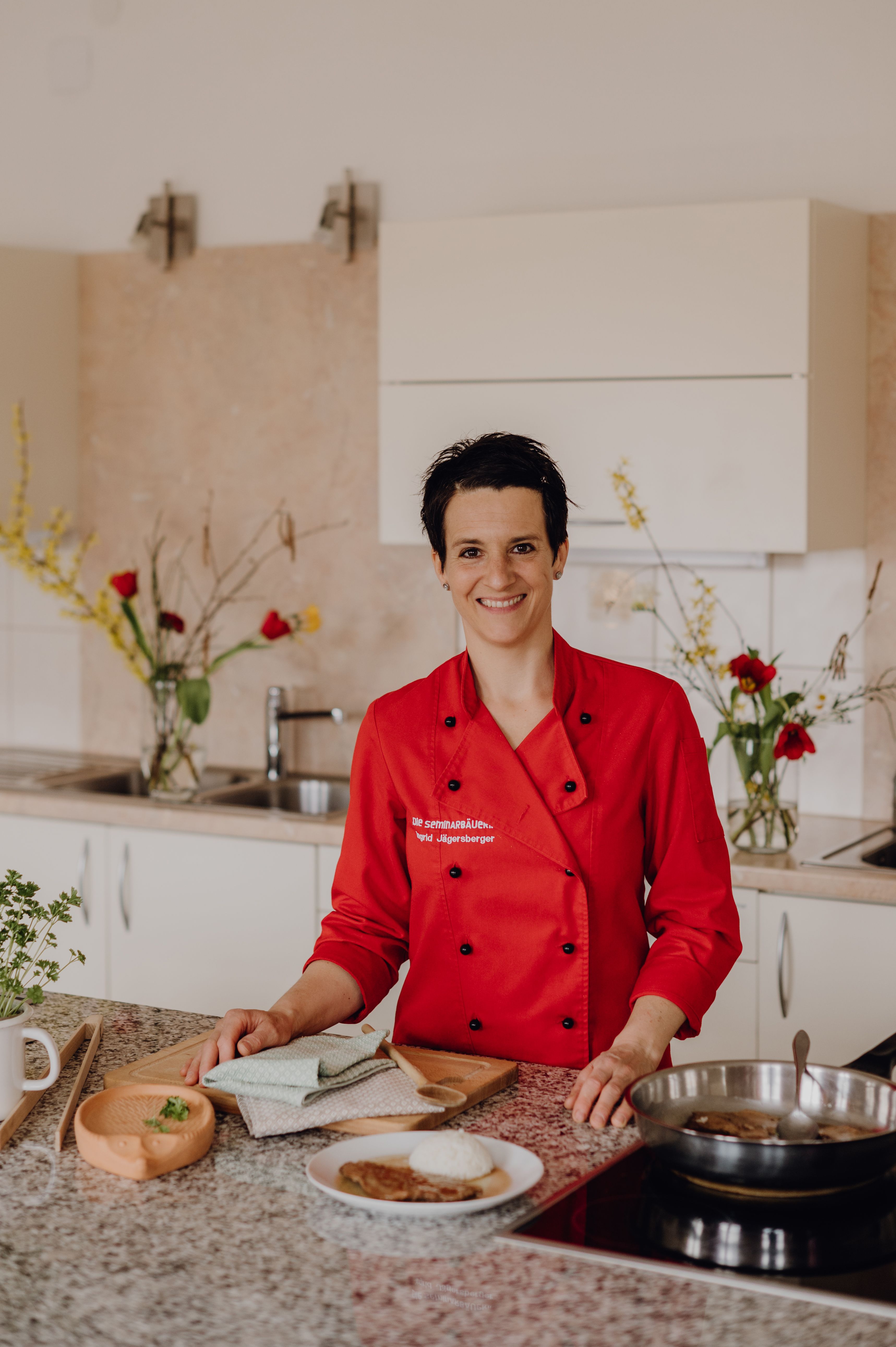 Eine Köchin in roter Uniform steht in einer Küche und lächelt in die Kamera. Vor ihr auf der Arbeitsplatte liegen Küchenutensilien und ein Teller mit Essen.