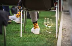 Person sitzt im Freien auf einer Veranstaltung; bunte Socken und zwei Weißweingläser stehen auf der Wiese neben den Stühlen.
