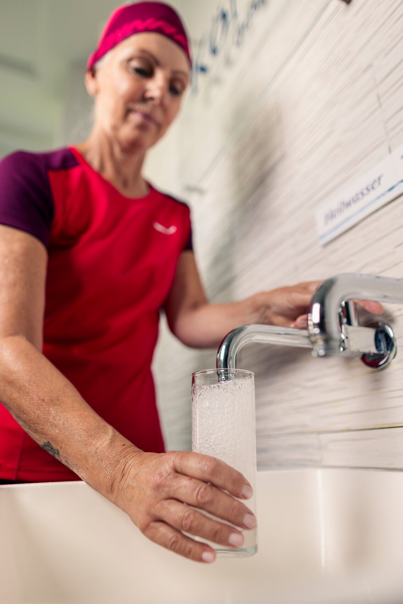 Eine sportlich gekleidete Person füllt an einem Wasserhahn ein Glas mit Wasser. 