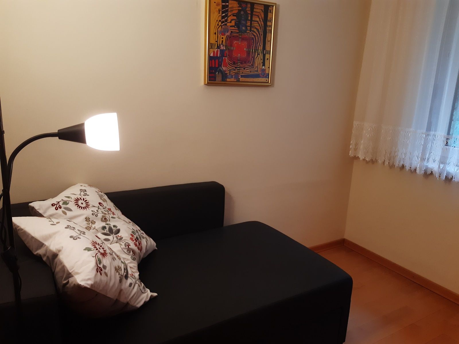 Gemütliche Leseecke mit schwarzem Sofa, Stehlampe und dekorativem Kissen. An der Wand hängt ein buntes Bild, daneben ein Fenster mit Gardine.