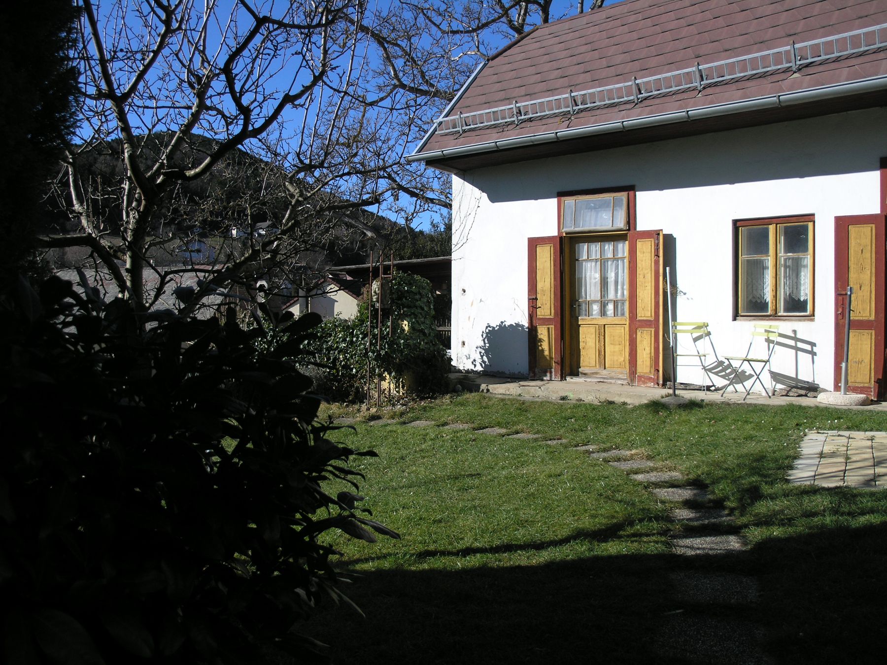 Ein Garten mit einem weißen Haus, braunen Türen und Fenstern. Im Vordergrund ein Baum und Gartenmöbel auf einer Terrasse.