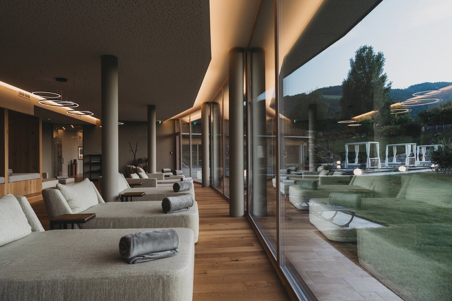 Ein eleganter Spa-Ruheraum mit warmem Dielenboden und runden Stützsäulen. Mehrere gepolsterte Daybeds mit grauen Decken sind ordentlich nebeneinander aufgereiht. Eine durchgehende Glasfront spiegelt den Innenraum und bietet gleichzeitig einen Ausblick in die dämmerige Natur.
