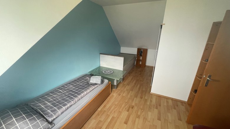 Ein Zimmer mit einem Einzelbett, einem kleinen Tisch und einem Regal. Die Wände sind blau und weiß gestrichen.