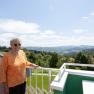 Ältere Frau mit Sonnenbrille steht auf einem Balkon mit Blick auf eine hügelige Landschaft.