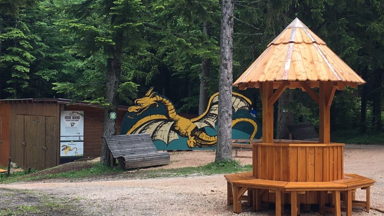 Ein hölzerner Pavillon auf einem Waldplatz mit einem gemalten Drachen im Hintergrund.