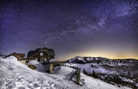Sternenklare Nacht über einer verschneiten Berghütte und Berglandschaft.