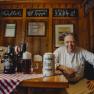 A landlord in traditional dress sits smiling at a table with beer mugs and bottles in a rustic dining room.