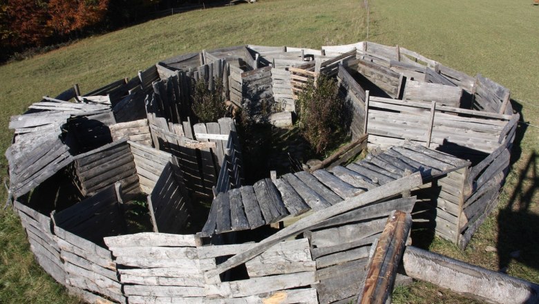 Ein aus Holzplanken gebautes Labyrinth auf einer Wiese, umgeben von Bäumen im Herbst.