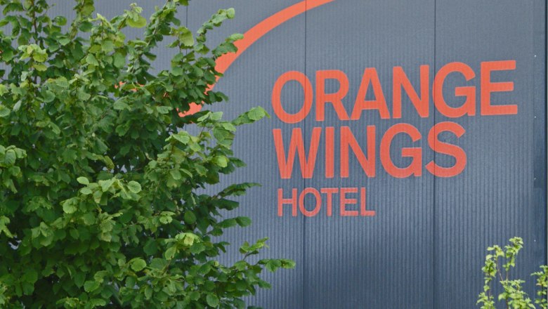Logo des Orange Wings Hotels auf einer Wand mit Pflanzen im Vordergrund.