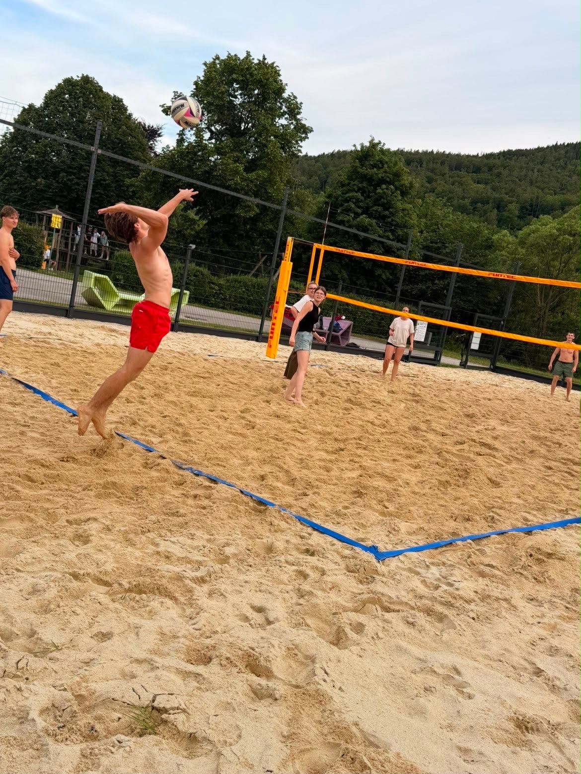 People playing beach volleyball on a sand court with trees in the background.