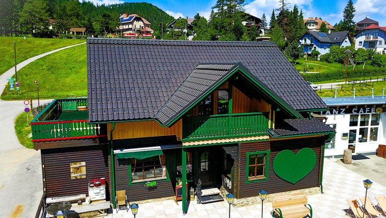 A traditional wooden house with a green heart motif and terrace in summer, surrounded by green hills and blue skies.