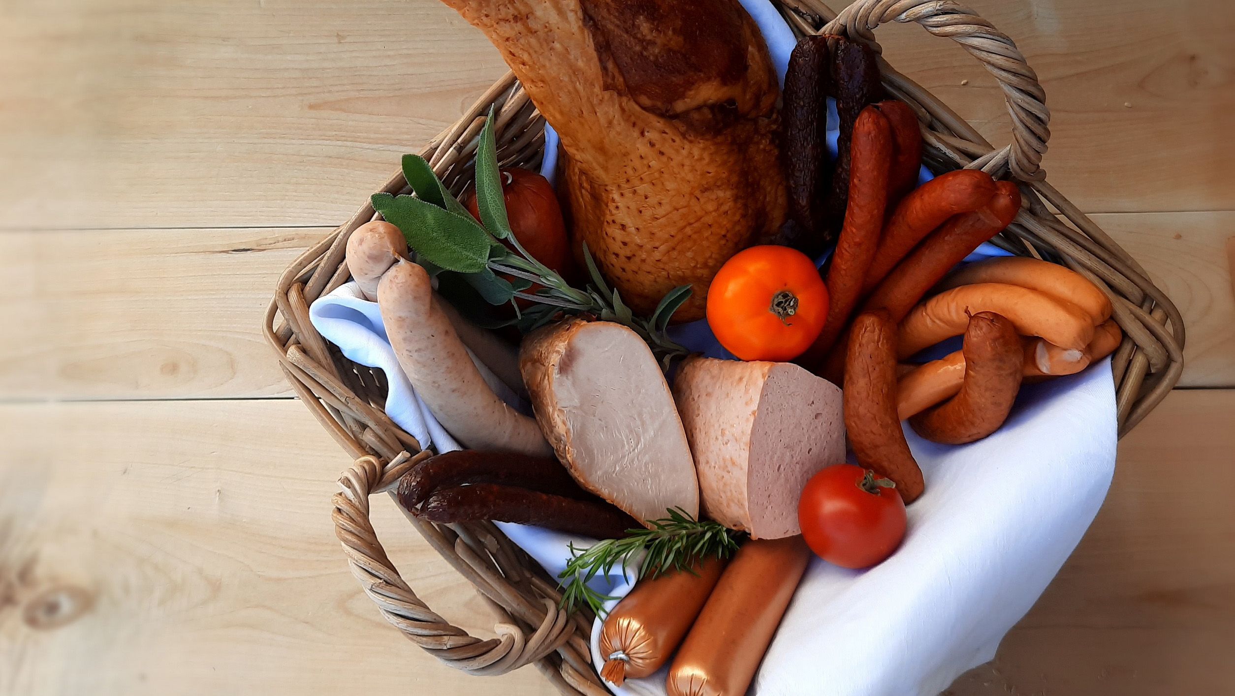 Diverse Wurstprodukte von der Bio-Pute in einem Korb mit Kräutern und Tomaten auf einem Holztisch.