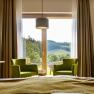 Interior view of a guest room with two green armchairs in front of a large window overlooking a hilly landscape.