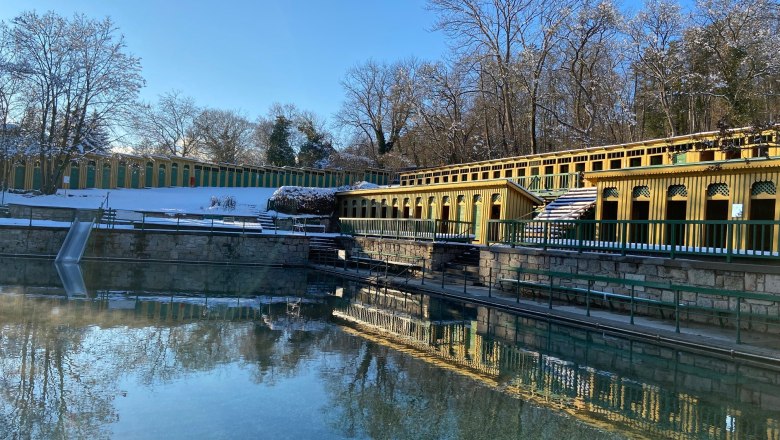 Außenansicht eines Thermalbads mit gelben Gebäuden und klarem Wasser, umgeben von schneebedeckten Bäumen.