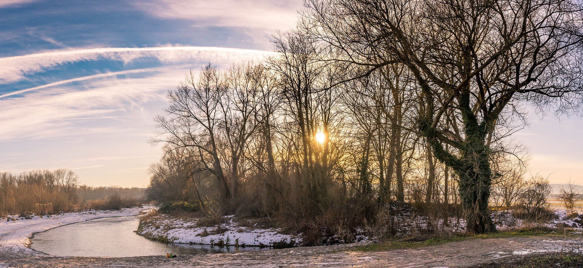 Bei einer Flussbiegung der Leitha mit teilweise schneebedecktem Ufer scheint die Sonne durch die kahlen Bäume.