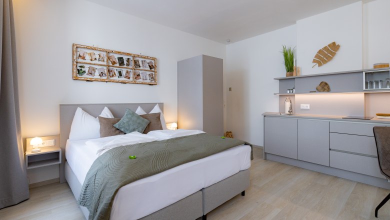 Modernes Schlafzimmer mit Doppelbett, grauer Bettwäsche, Wanddekoration und kleiner Küchenzeile.