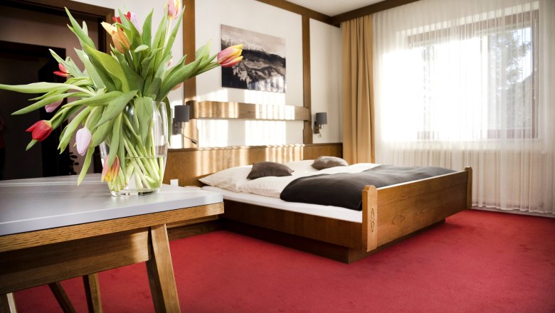 Ein gemütliches Doppelzimmer mit einem Holzbett, roten Teppichboden und einem Tisch mit einem Blumenstrauß aus Tulpen.