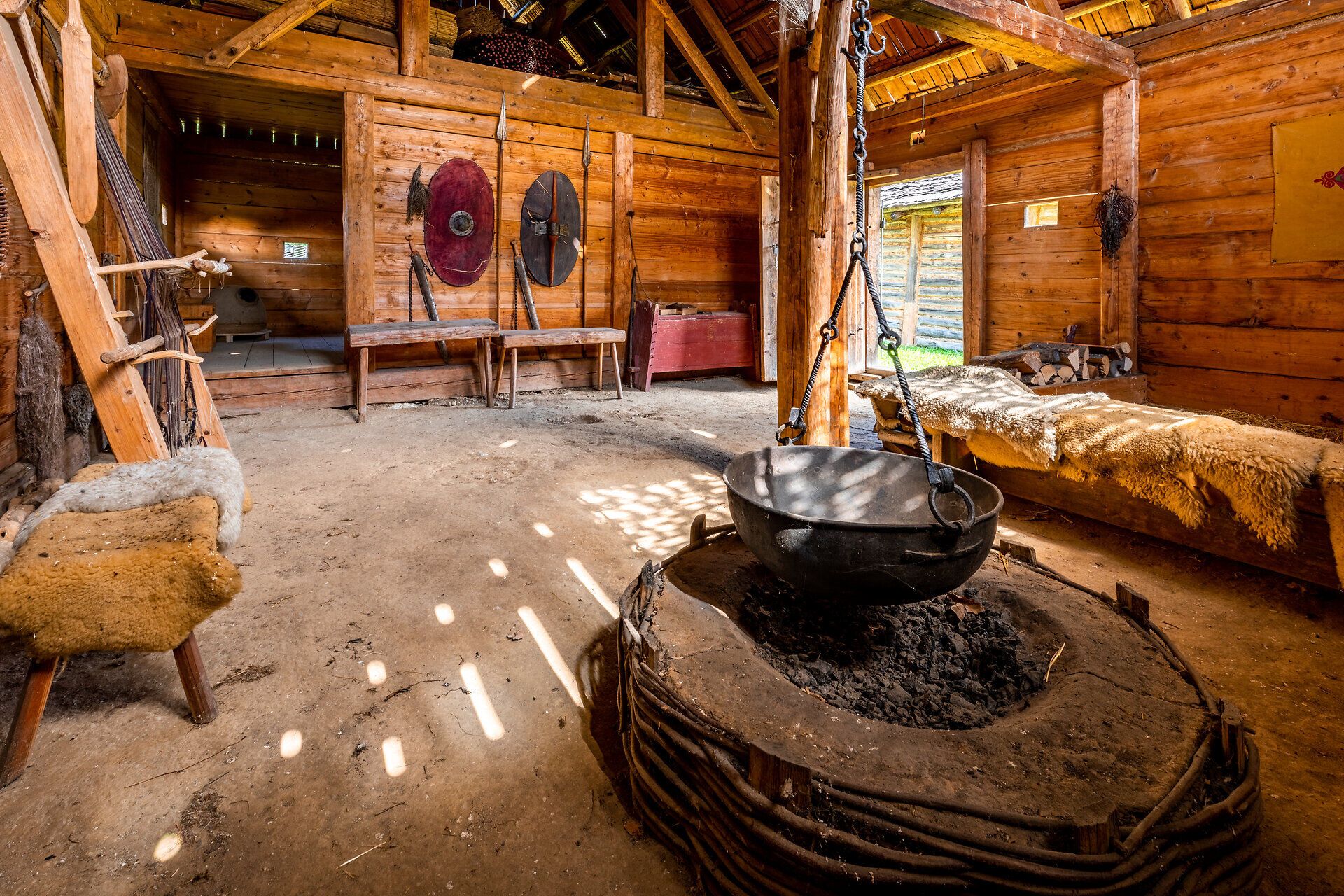 Innenansicht eines Hauses im Keltendorf Schwarzenbach mit Einrichtung. Im Zentrum des Raumes hängt ein Kessel über einer Feuerstelle. 