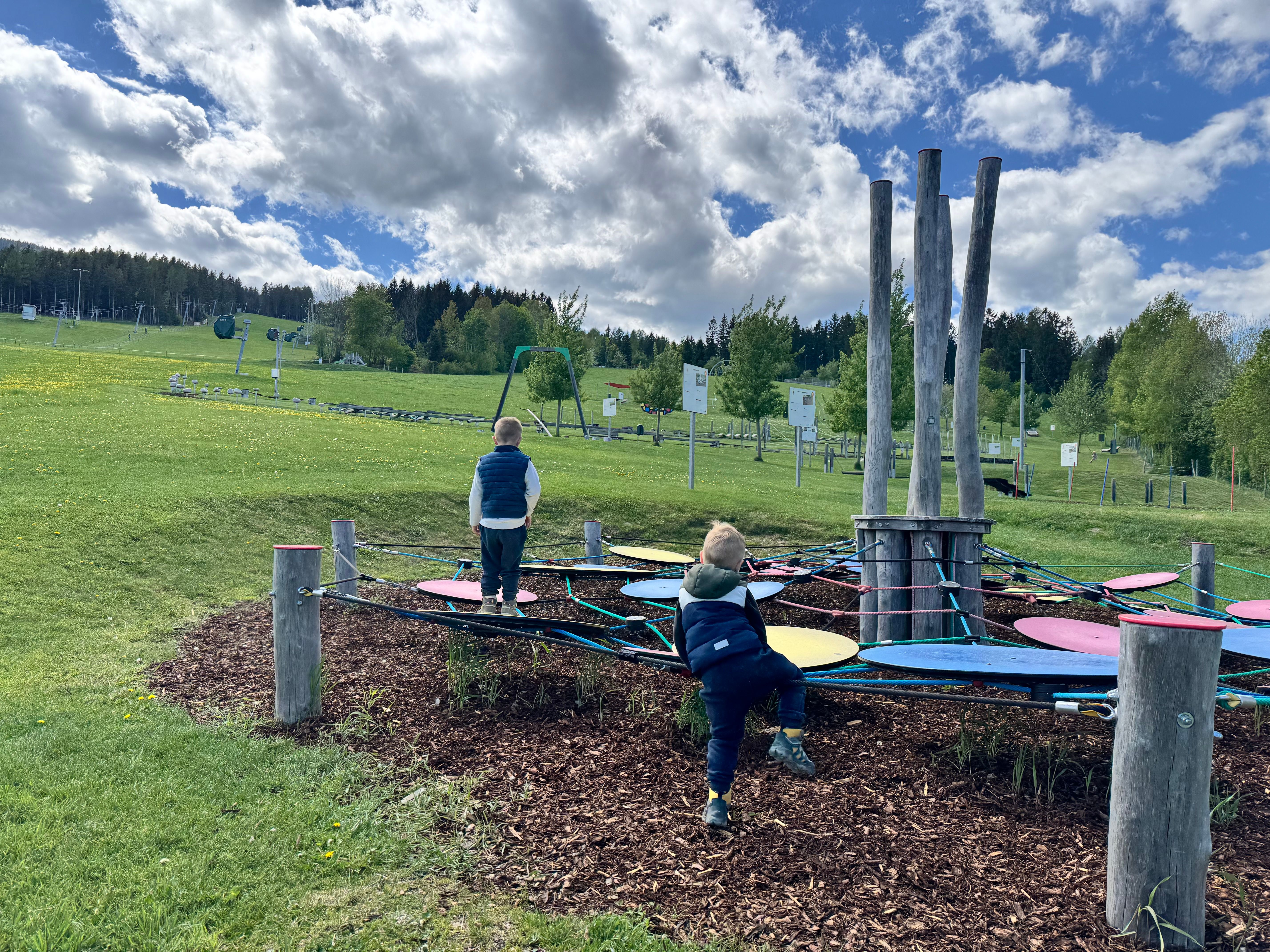 Zwei Kinder spielen auf einem Klettergerüst in einer grünen Landschaft mit Bäumen und Wolken am Himmel.