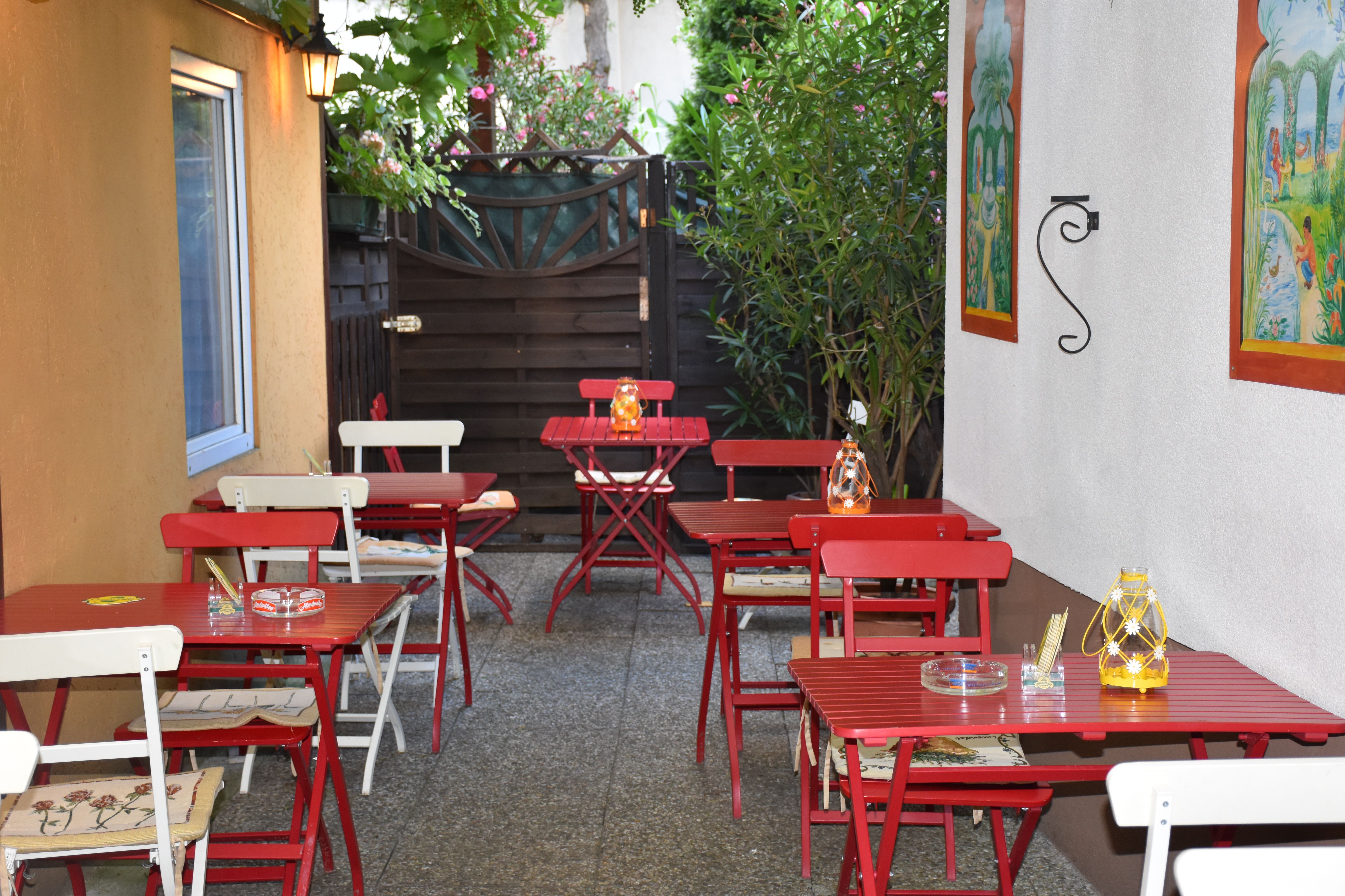 Ein gemütlicher Schanigarten mit roten Tischen und Stühlen, dekoriert mit Laternen und Pflanzen.