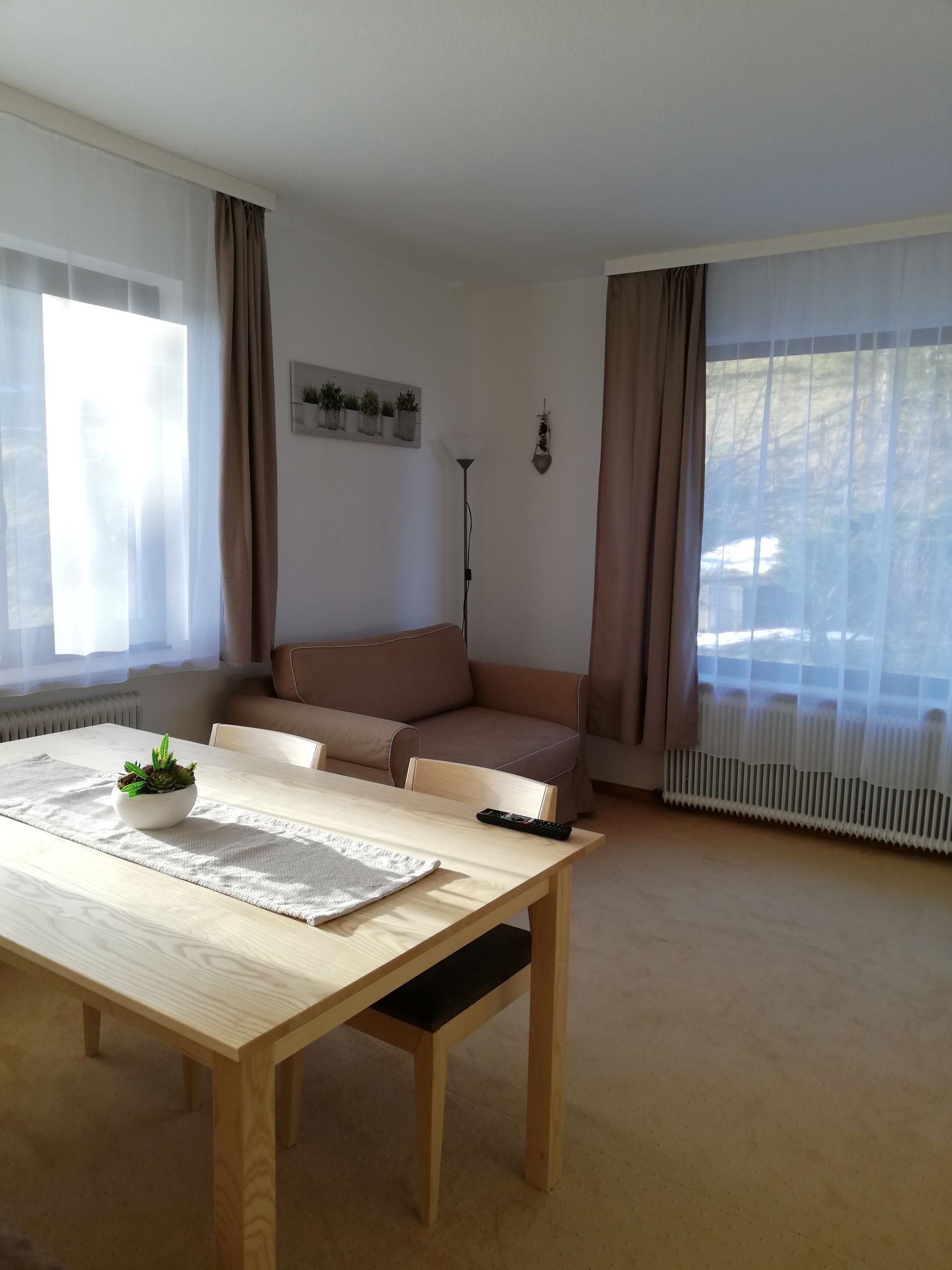 Helles Wohnzimmer mit Esstisch, Stühlen, Sofa und großen Fenstern mit Vorhängen.
