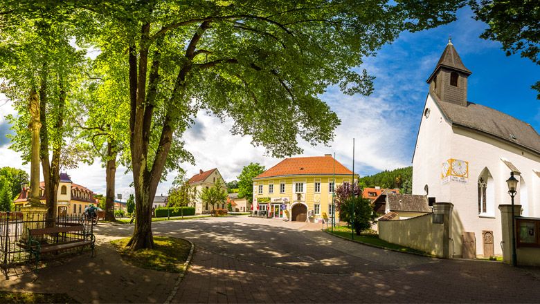 Panoramablick auf den Kirchenplatz von Feistritz mit Kirche, Bäumen und historischen Gebäuden.