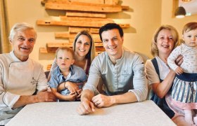 A family sits at a table and smiles at the camera. Two adults are each holding a child on their lap.