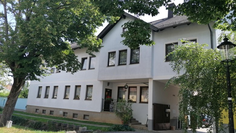 Außenansicht eines weißen Gebäudes mit vielen Fenstern, umgeben von Bäumen.