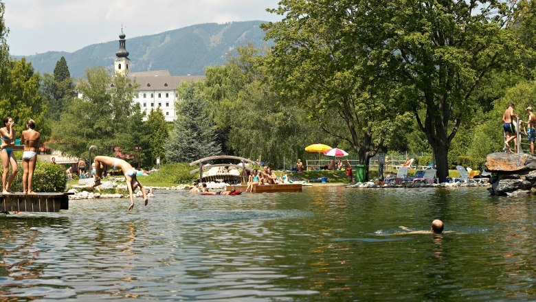 Menschen genießen einen sonnigen Tag im Naturbad Gloggnitz mit Schwimmen und Sonnenbaden, umgeben von Bäumen und Bergen.