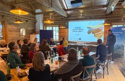 Workshop Teilnehmer sitzen in einem rustikalen Raum an Tischen, ein Mann moderiert den Workshop vor den Teilnehmern vor einer Leinwand. 