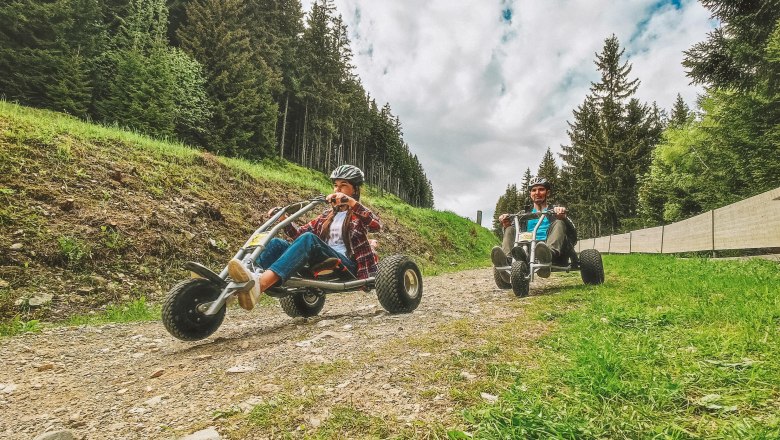 Zwei Personen fahren mit Mountaincarts auf einem Schotterweg im Wald.