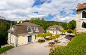 Zwei Kavaliershäuser mit Mansardendächern und gepflegten Bäumen im Vordergrund, umgeben von grüner Landschaft und blauem Himmel.