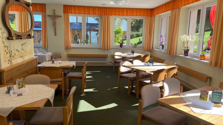 Ein heller Speisesaal mit Holztischen, St&uuml;hlen und gro&szlig;en Fenstern mit orangefarbenen Vorh&auml;ngen.