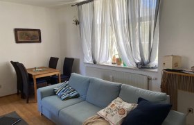 Ein Wohnzimmer mit einem blauen Sofa, einem Holztisch mit St&uuml;hlen und einem Fenster mit wei&szlig;en Vorh&auml;ngen.