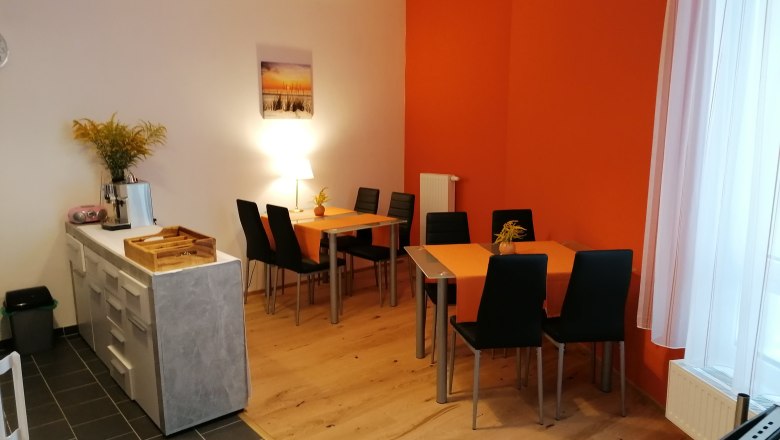 A breakfast room with two tables, black chairs and orange tablecloths. A chest of drawers with a coffee machine is on the left.