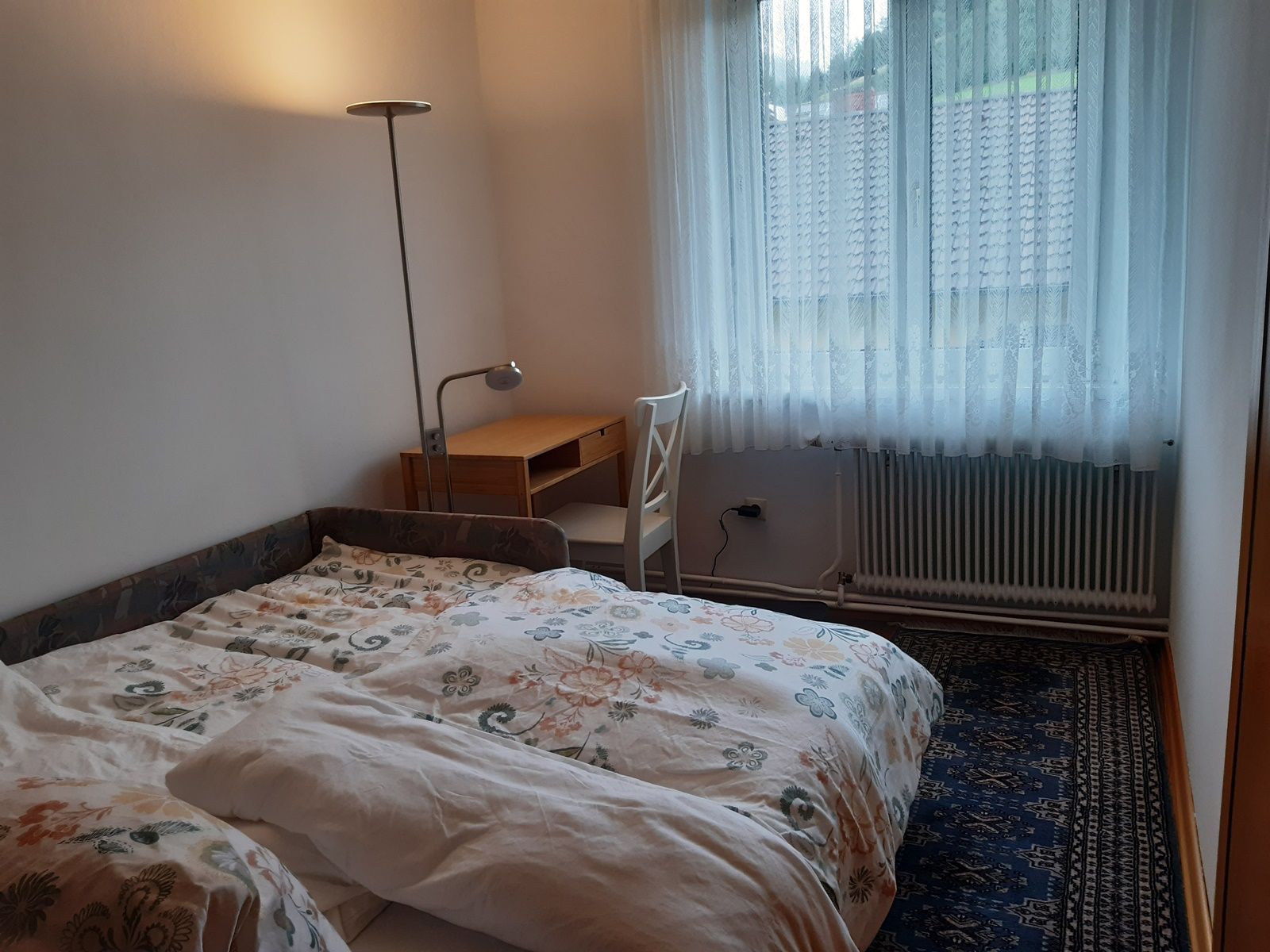 Ein Zimmer mit einer ausgezogenen Couch als Bett, einem Schreibtisch mit Stuhl und einem Fenster mit Vorhängen.