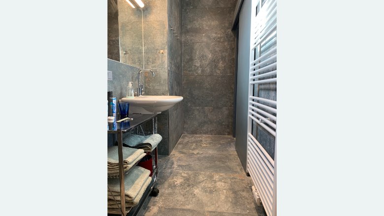 Modernes Badezimmer mit grauen Fliesen, Waschbecken, Handtuchregal und Heizk&ouml;rper.