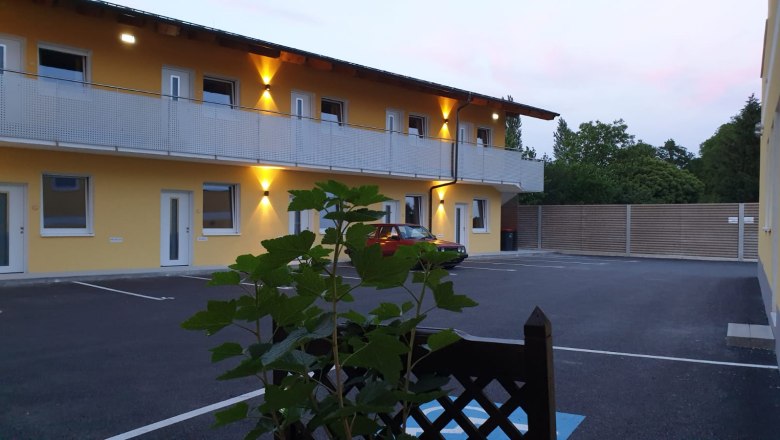 Aussenansicht, © Alexandra Heiling-Niegl Außenansicht eines zweistöckigen Gebäudes mit gelber Fassade und beleuchtetem Balkon, davor ein geparkter roter Wagen und Pflanzen im Vordergrund.