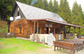 A wooden hut in the forest with tables and benches in front of it.