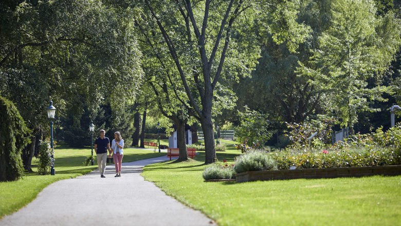 Ein Paar spaziert auf einem Weg im Kurpark Bad Schönau, umgeben von grünen Bäumen und gepflegten Blumenbeeten.