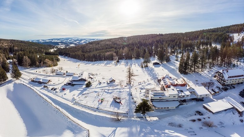 Luftaufnahme eines verschneiten Skigebiets mit Gebäuden und Wald im Hintergrund.