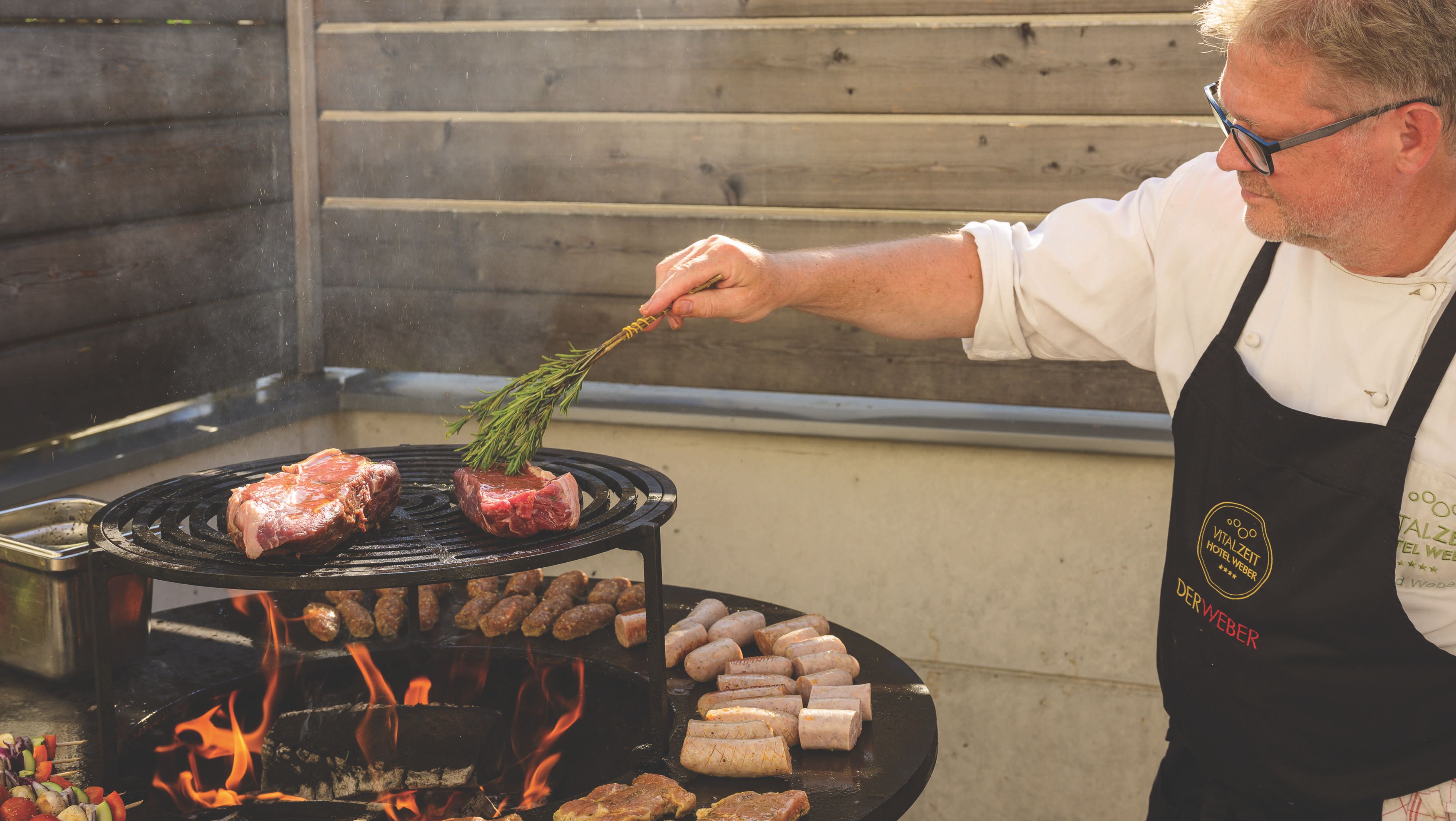 Der Chef des Hauses grillt Fleisch und Würstchen auf einem runden Grill mit offenem Feuer.