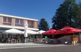 Ein moderner Gastgarten mit Sonnenschirmen und Sitzgelegenheiten vor einem Gebäude bei sonnigem Wetter.