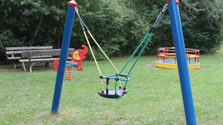 Spielplatz mit Schaukel, Wippe und Karussell auf einer Wiese.